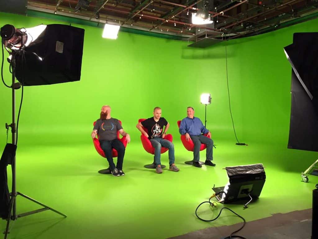 Three men on set sitting in red chairs against a green screen